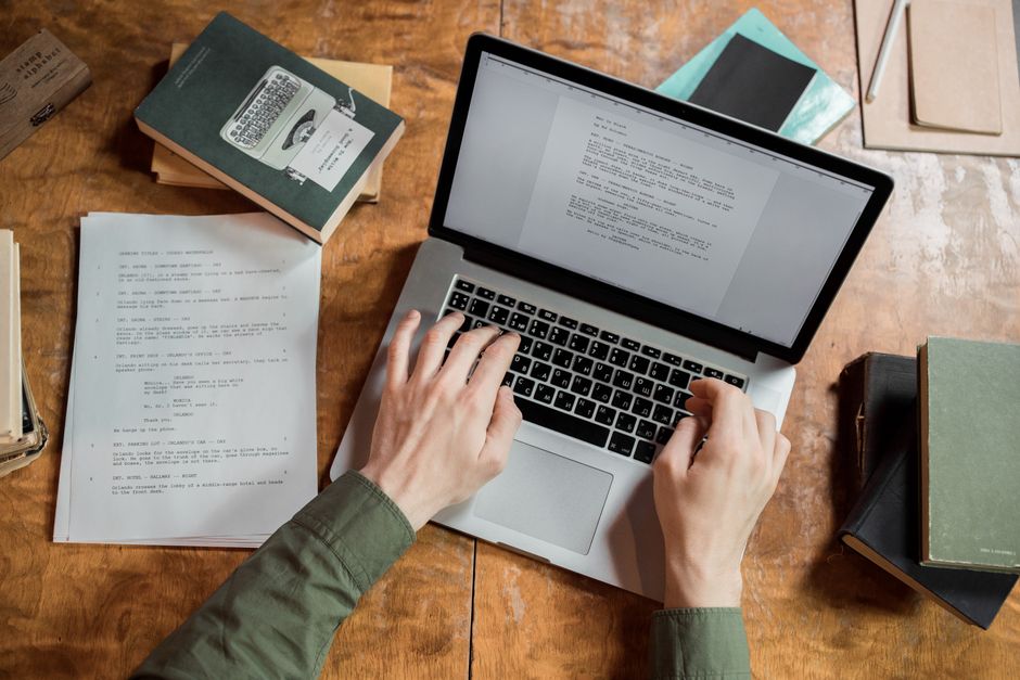 Mani che digitano su un laptop su un tavolo di legno con alcuni libri sparsi intorno.